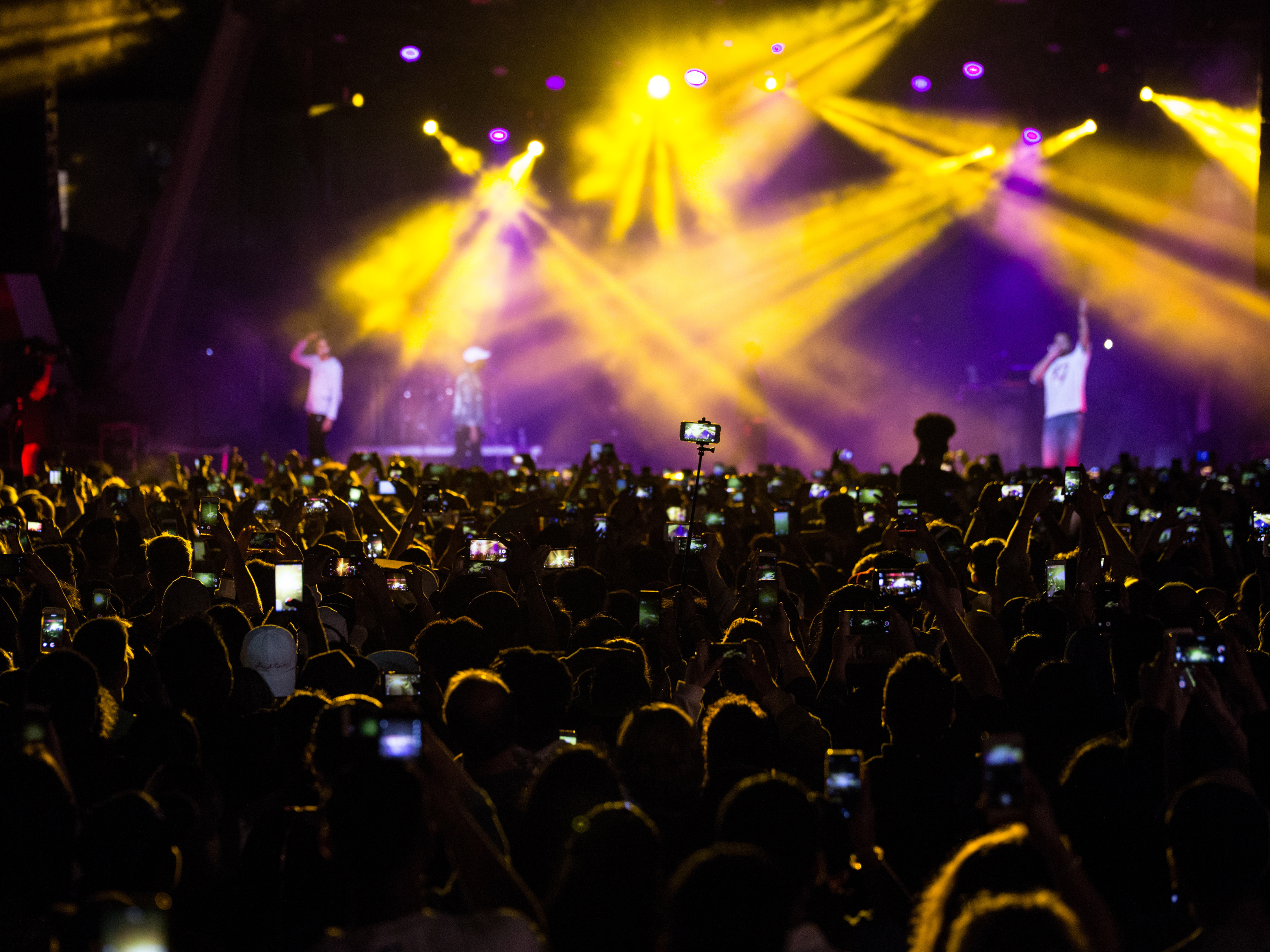 Concert crowd raising phones toward two performers on stage lit by yellow and purple spotlights